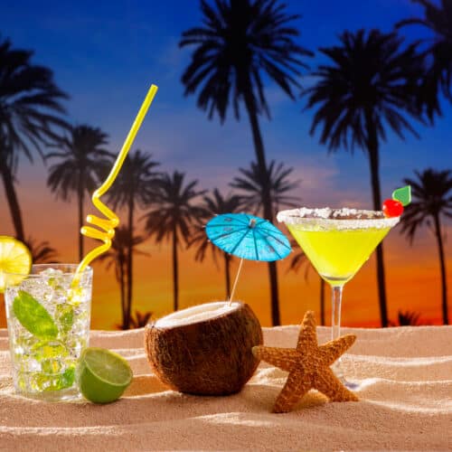 Three cocktails including one in a coconut with an umbrella on a sandy beach at sunset