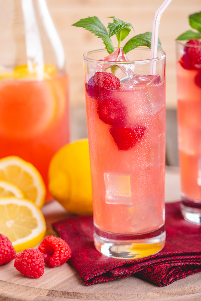 Closeup of a refreshing glass of raspberry lemonade with fresh berries and lemon in background