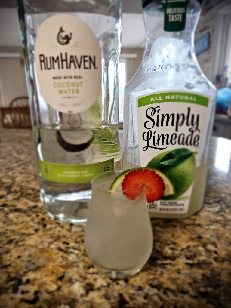 Ingredients for RumHaven Limeade Cocktail: Bottles of Rum Haven Caribbean Rum, Simply Limeade and a small glass with mixture and strawberry garnish