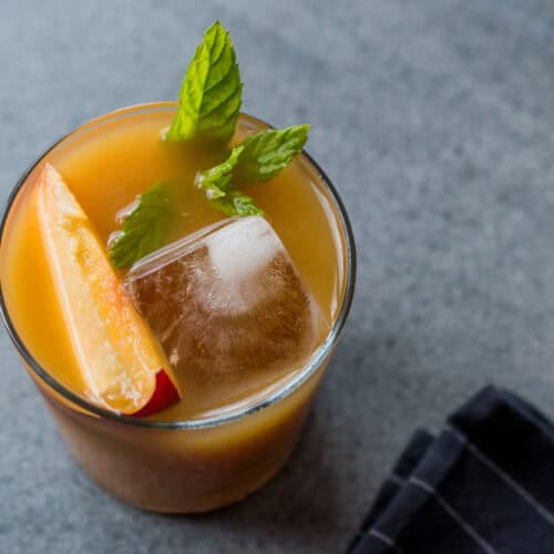 A bourbon glass with one large ice cube and slice of peach and mint.