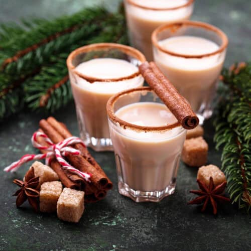 4 shot glasses with eggnog inside and a cinnamon stick on top with Christmas decorations