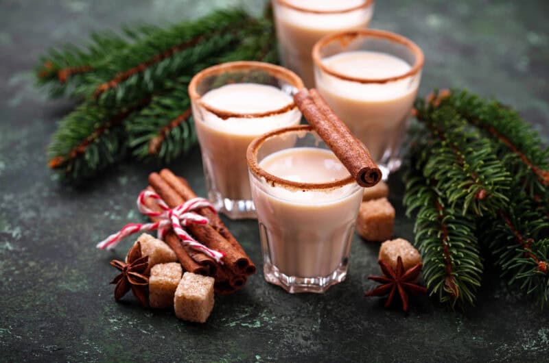 4 shot glasses with eggnog inside and a cinnamon stick on top with Christmas decorations