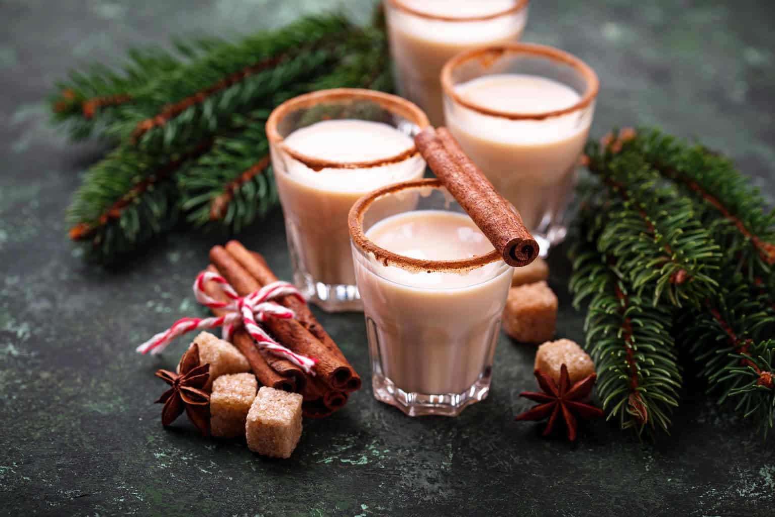 4 shot glasses with eggnog inside and a cinnamon stick on top with Christmas decorations