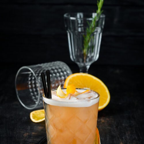 Whikey sour cocktail in a clear glass with a foamy egg white top and orange peel garnish