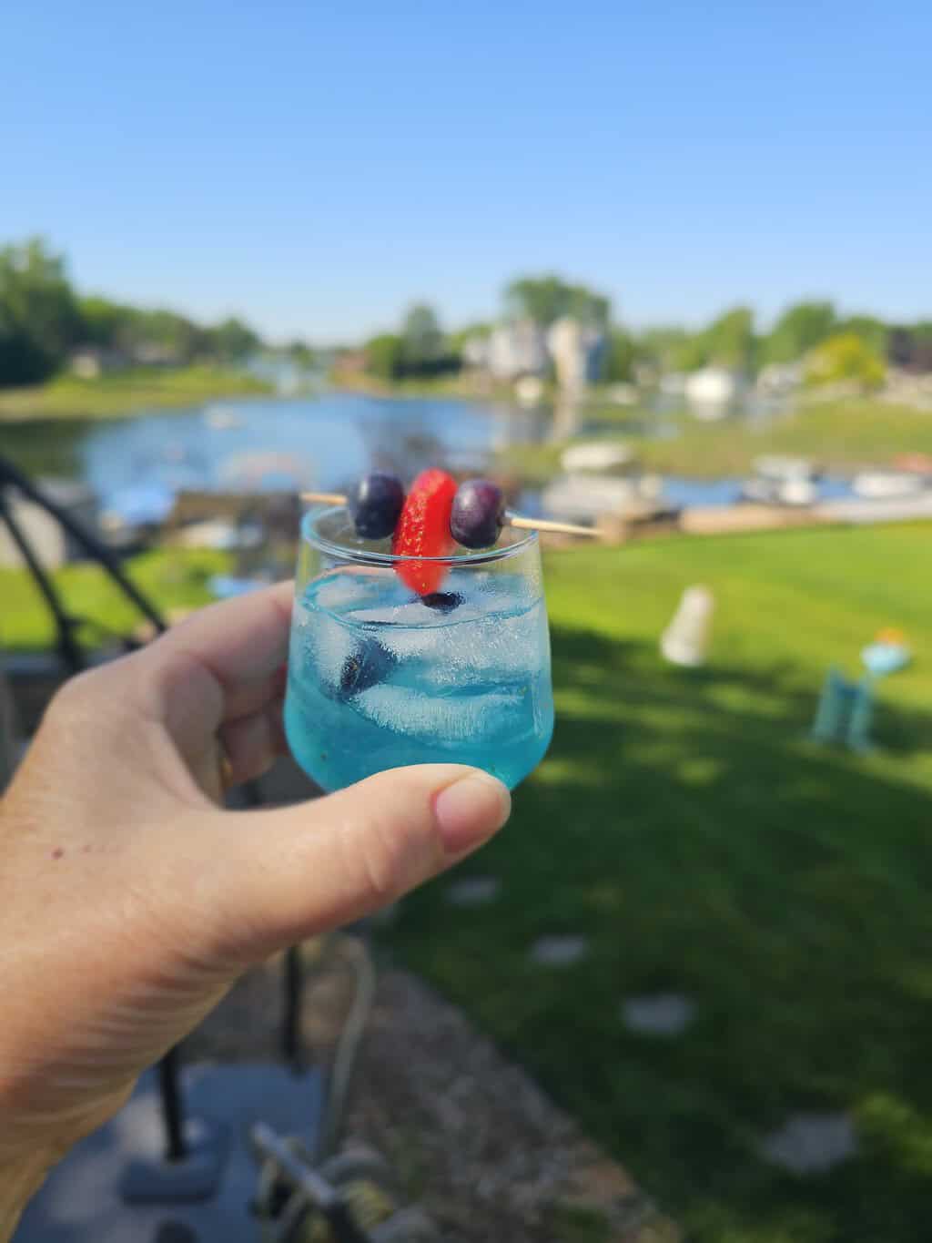 Hand holding a blue cocktail with ice, garnished with olives and a red pepper on a skewer, in an outdoor setting with a blurred lawn and water in the background.