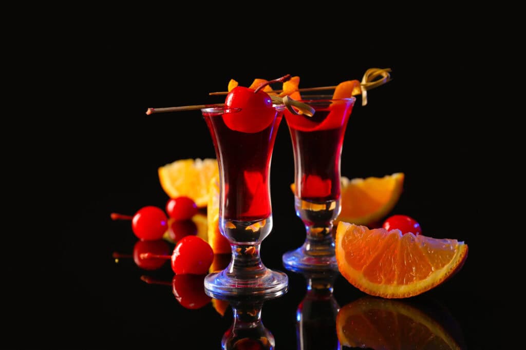 Two red cocktails in tall glasses with cherry skewers and orange wedges on a glossy black surface for Pregaming at home