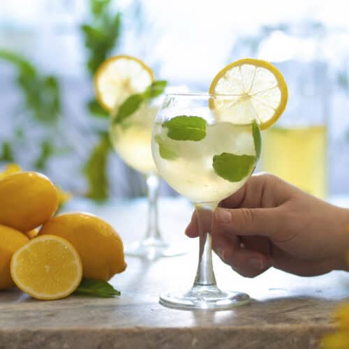 Hand holding a glass of limoncello Hugo Sprtz with mint with a lemon garnish, foreground on a table with lemons and yellow flowers in background